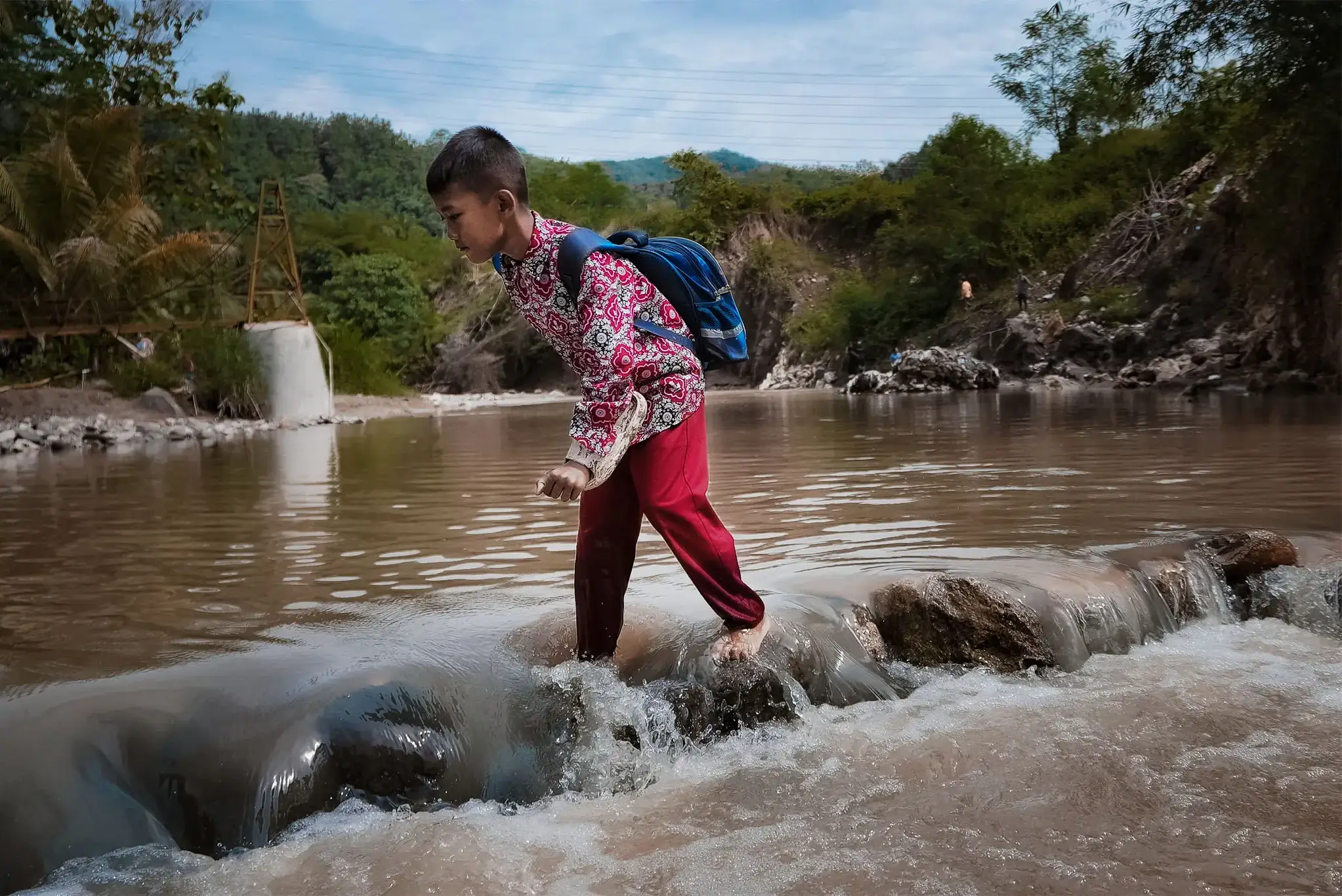 A child in colorful clothing balances on rocks in a shallow river.
