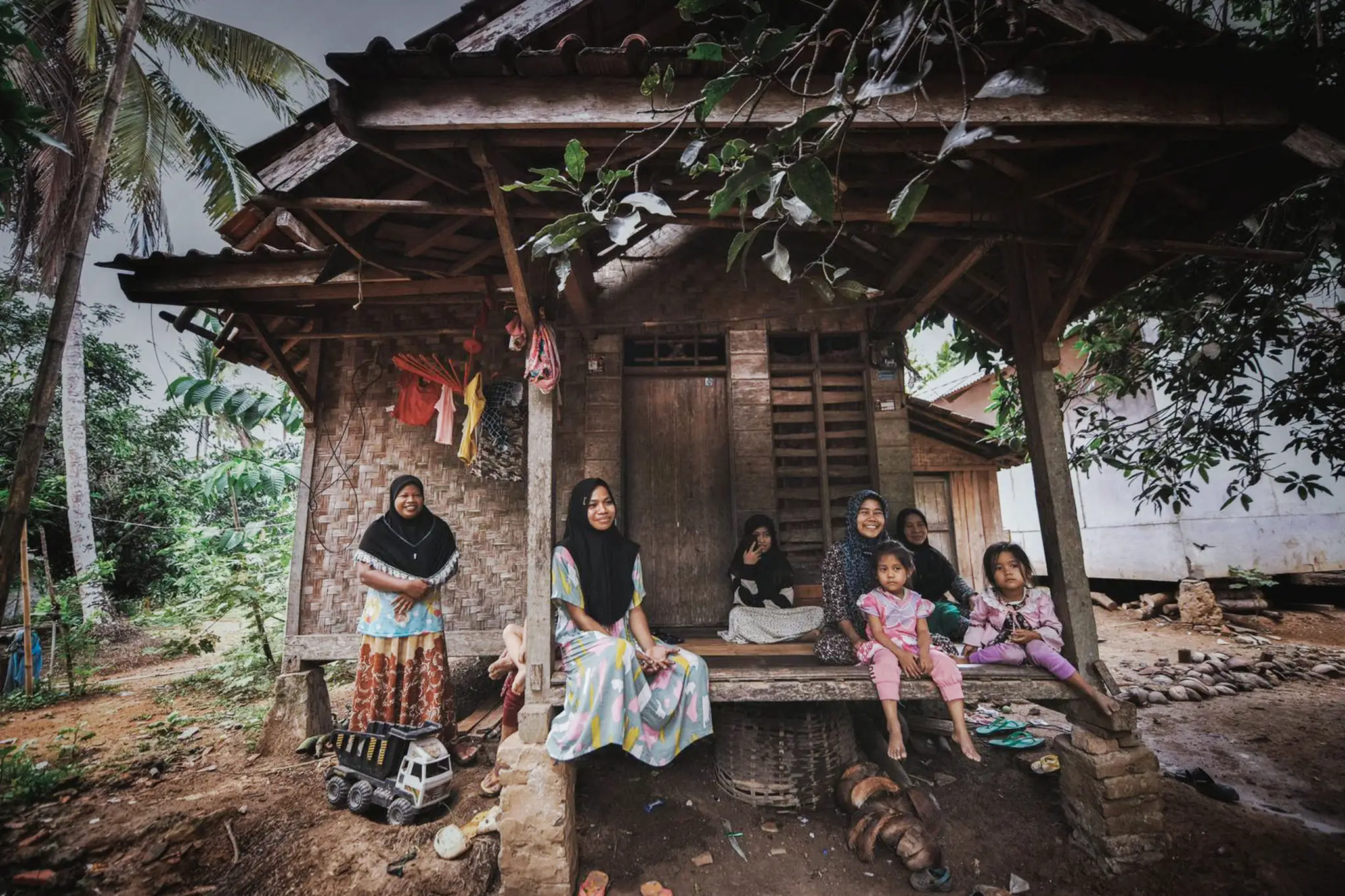 A group of women and children relax on the porch of a rustic wooden house surrounded by greenery and palm trees.
