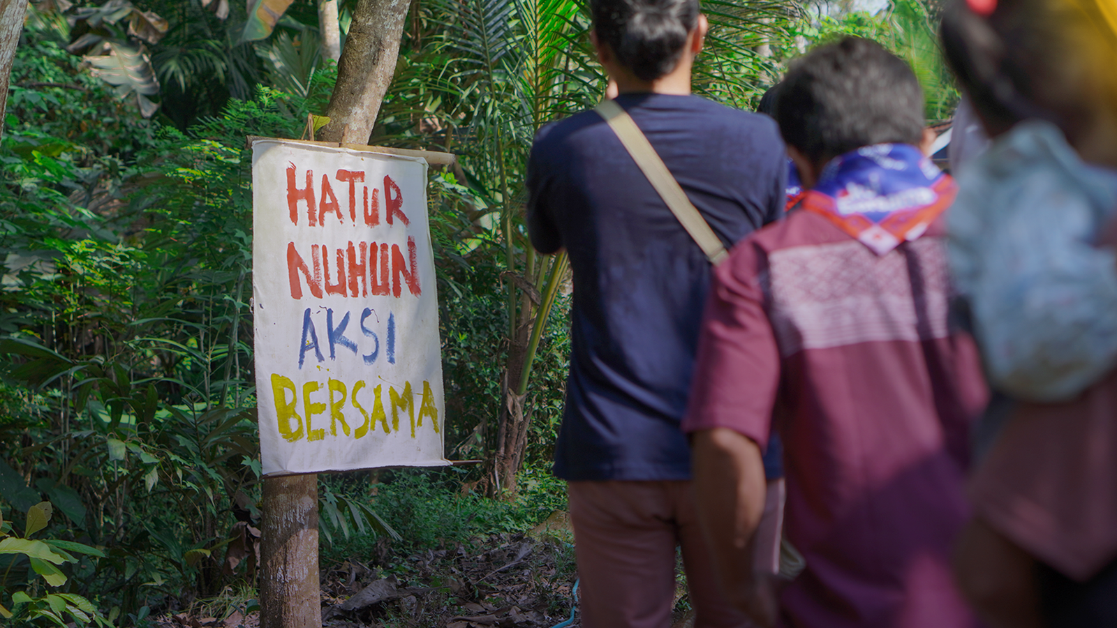 Sekelompok orang berjalan menuju tanda warna-warni bertuliskan "Hatur Nuhun Aksi Bersama" di area luar ruangan yang hijau dan rindang.
